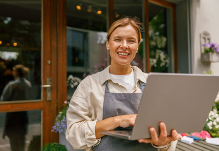 Smiling woman in a work apron with a laptop standing outside a business