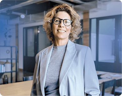 Middle aged woman in a gray business suit and wearing glasses smiling inside an office setting